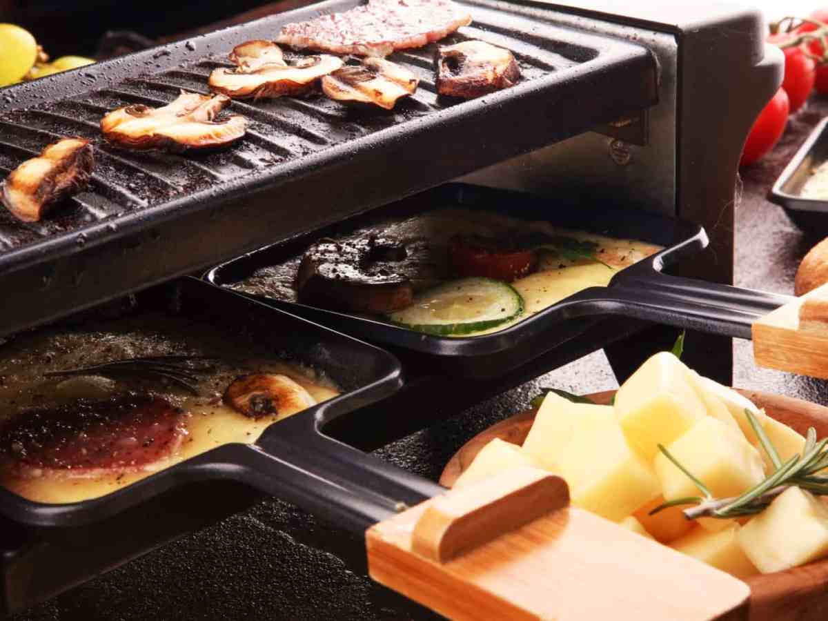 Nahaufnahme eines Raclette-Grills, auf der Grillplatte liegen Pilze, die Pfännchen sind befüllt und mit Käse überdeckt