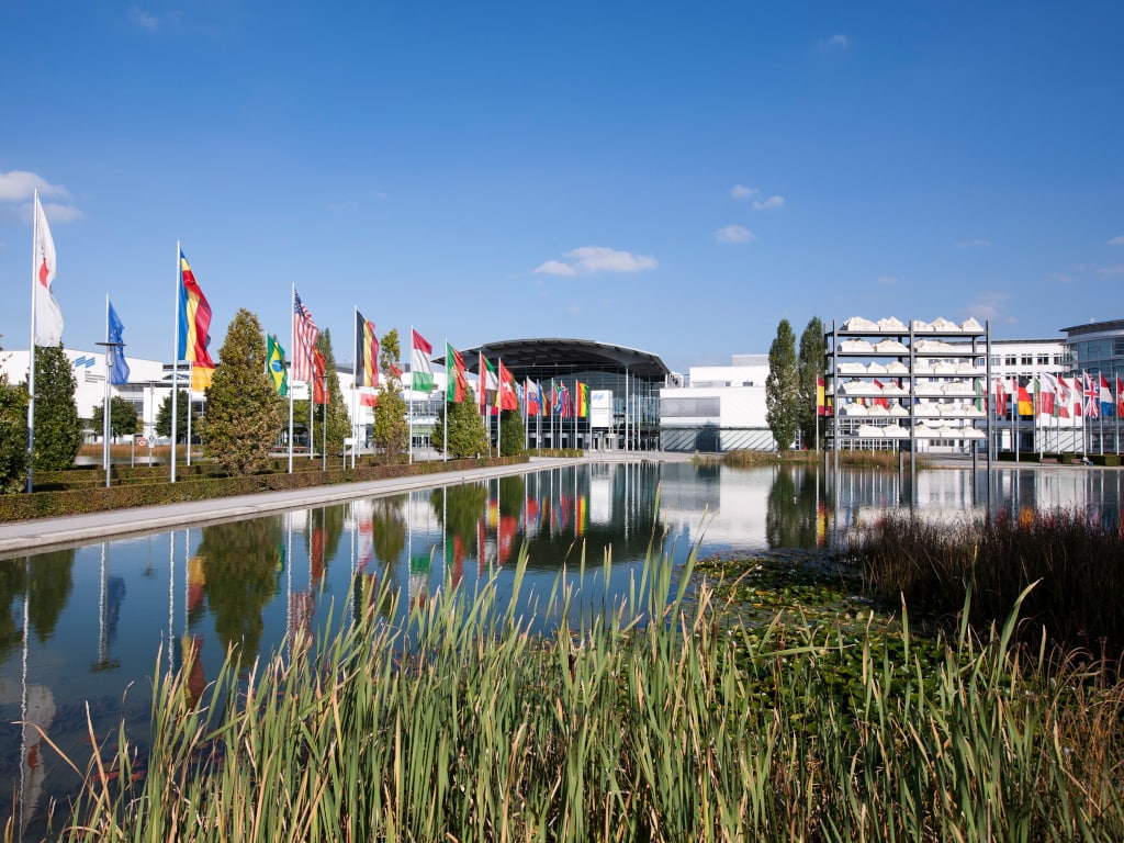 Großes langes Wasserbecken mit verschiedenen Fahnenmasten auf der linken Seite und dem Messegebäude in München im Hintergrund