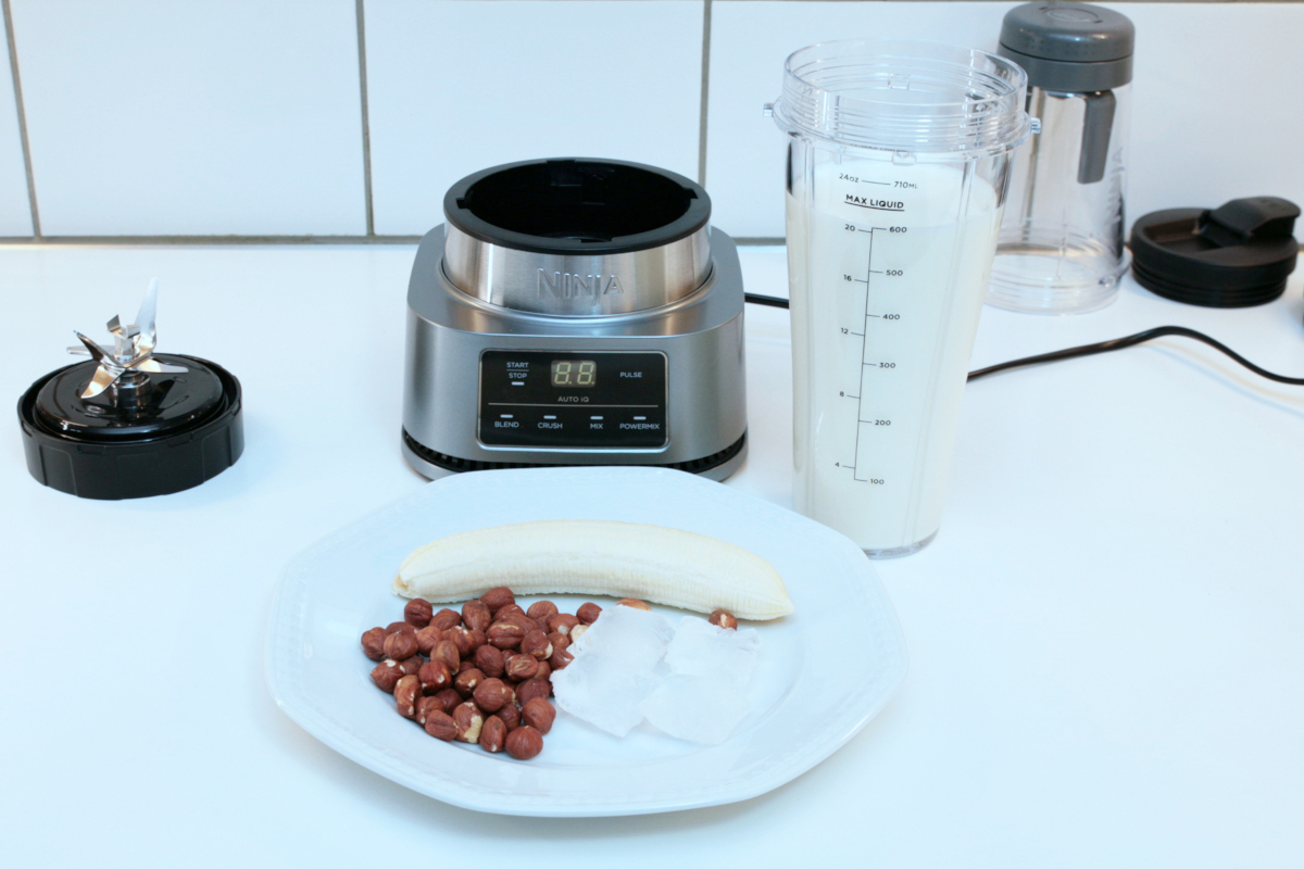 Teller mit Banane und Haselnüssen vor silbernem Standmixer, daneben Becher mit weißem Smoothie