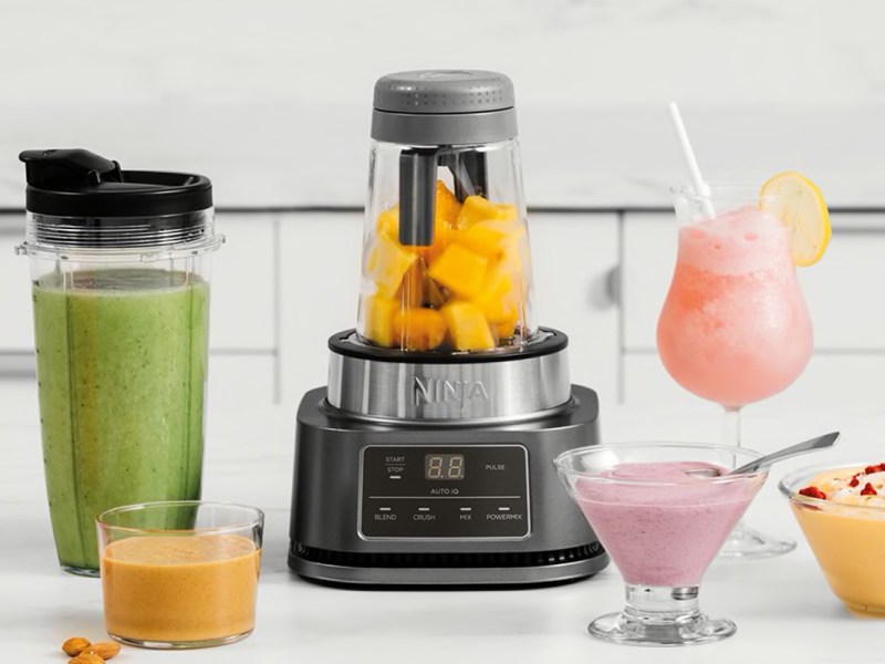 Grauer Standmixer in der Mitte mit gelben Stücken Obst, daneben verschiedene Becher, Gläser und schüsseln mit unterschiedlich farbigen Smoothies, Saucen und Getränke