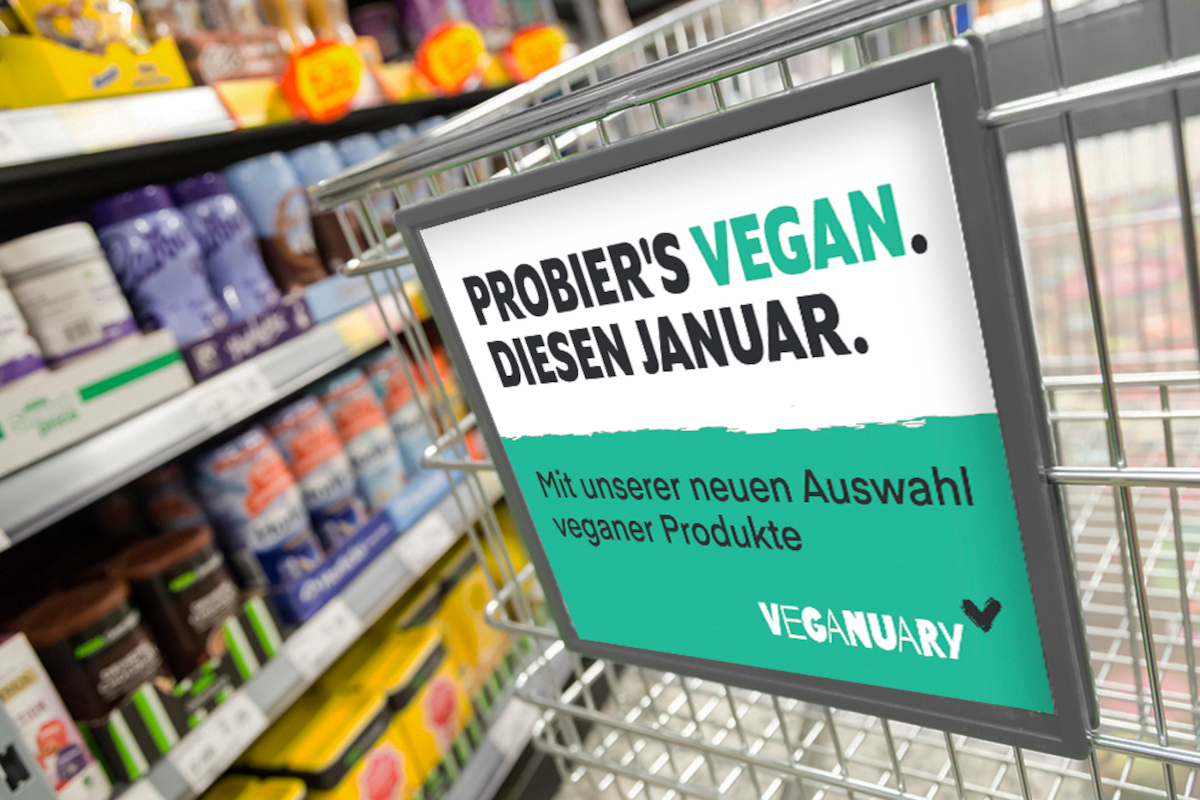 Ecke Einkaufswagen mit grün weißem schild vorne dran von Veganuary, im Hintergrund ein Regal im Supermarkt