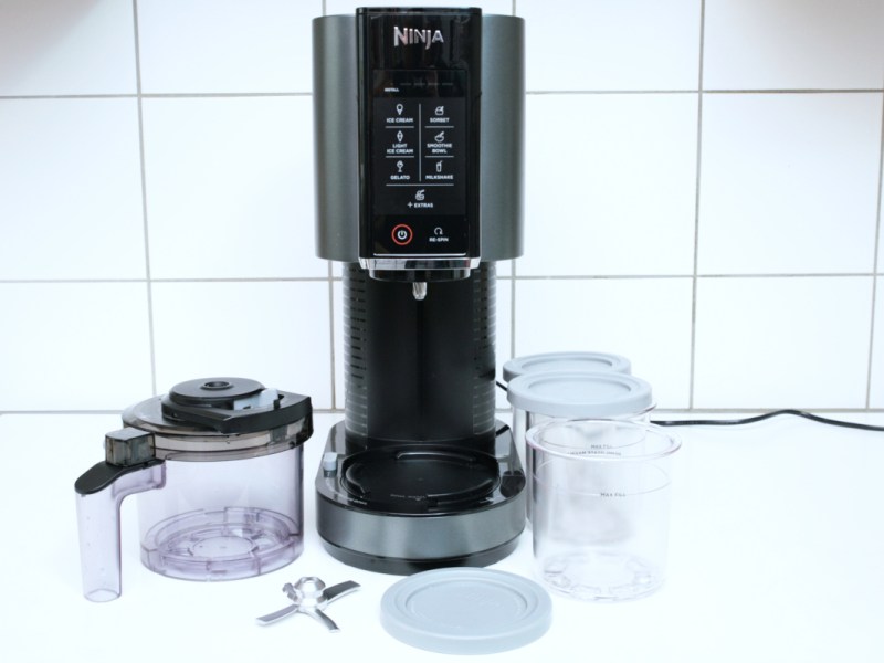 Silbern schwarze Ninja-Eismaschine auf weißer Arbeitsfläche von vorne mit Behältern und Klinge daneben