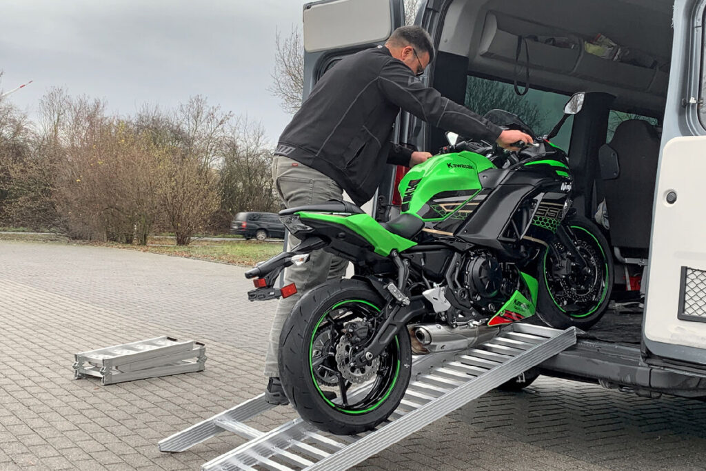 Mann schiebt Motorrad über eine Auffahrrampe in einen Kleintransporter.