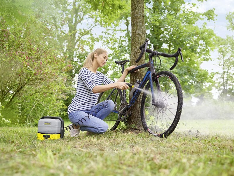 Frau mit blinden langen Haaren in gestreiftem Shirt und Jeans kniet vor dreckigem blauen Rennrad und hält Pistole von Kärcher Niederdruckreiniger auf Wiese mit Bäumen im Hintergrund