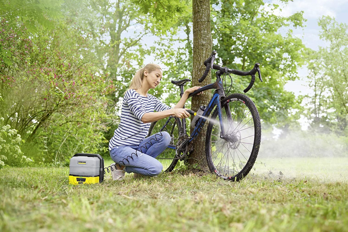 Frau mit blinden langen Haaren in gestreiftem Shirt und Jeans kniet vor dreckigem blauen Rennrad und hält Pistole von Kärcher Niederdruckreiniger auf Wiese mit Bäumen im Hintergrund