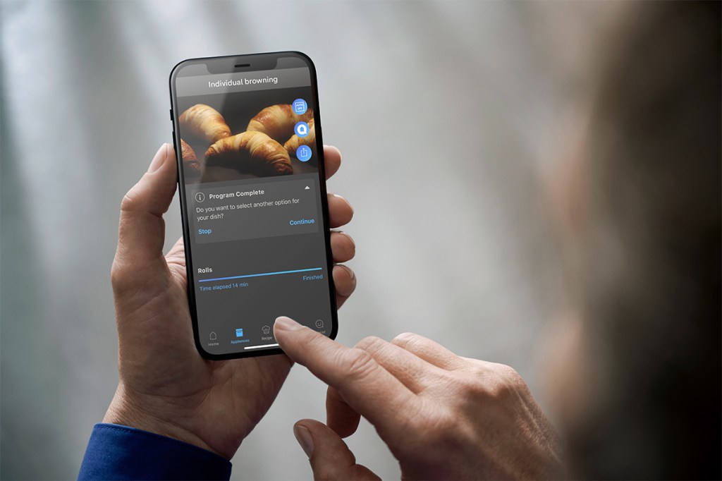 Es ist eine Person zu sehen, die am Smartphone die App für den neuen Siemens iQ700 bedient. Auf dem Display sieht man ansprechend gebräunte Croissants.