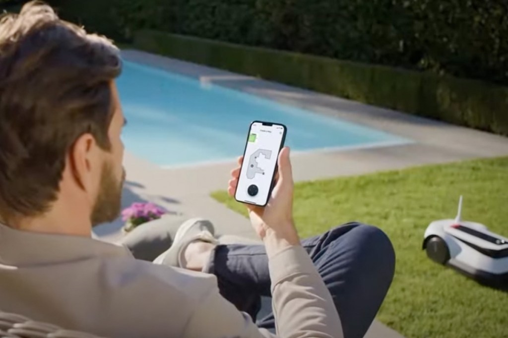 Eine Person schaut aufs Handy mit der Ecovacs-App. Im Hintergrund fährt der neue Mähroboter.