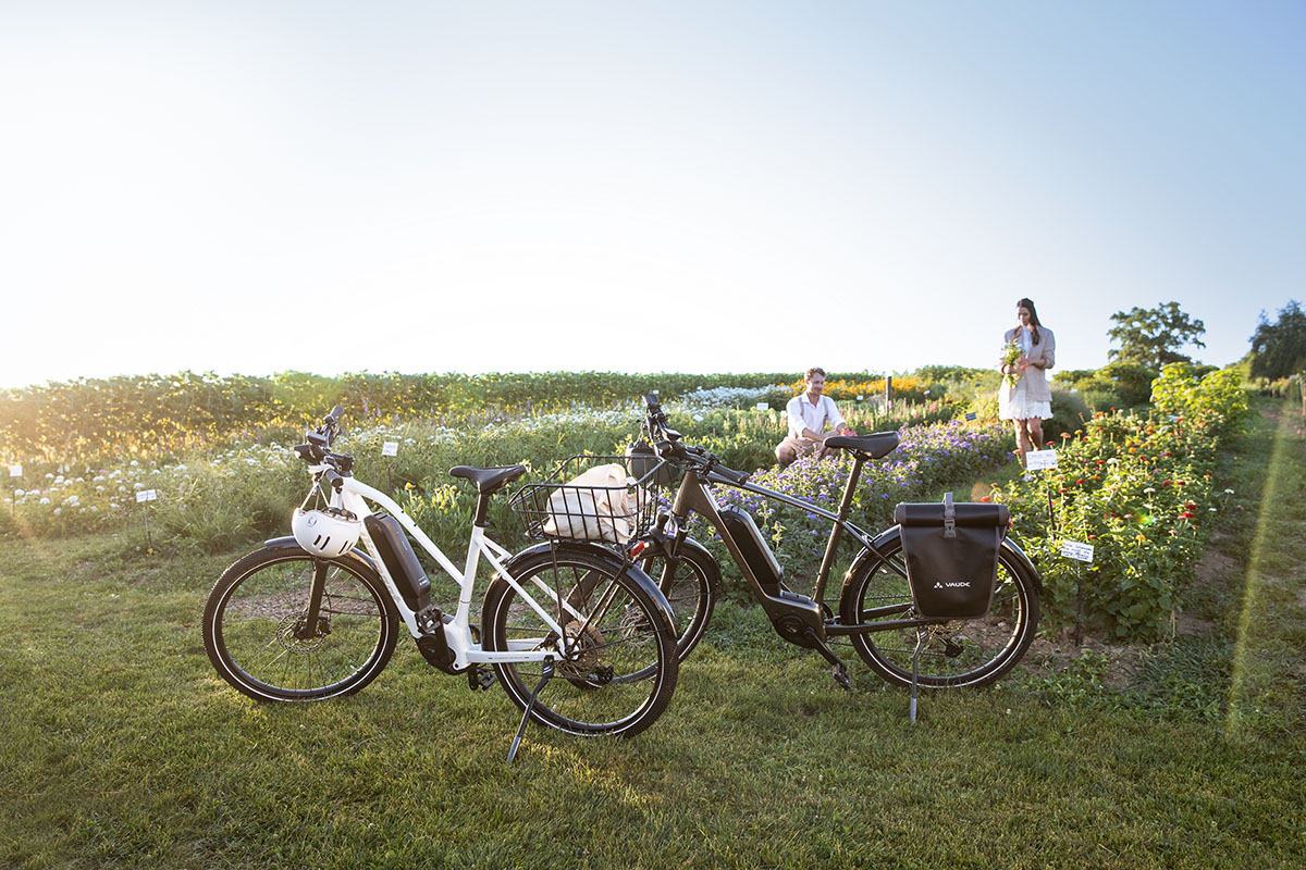 E-Bike-Neuheiten Diamant Mandara 160 & Zing CHC: wei E-Bikes stehen auf einer Wiese, dahinter ein Fraun und Mann in einem Blumenbeet.