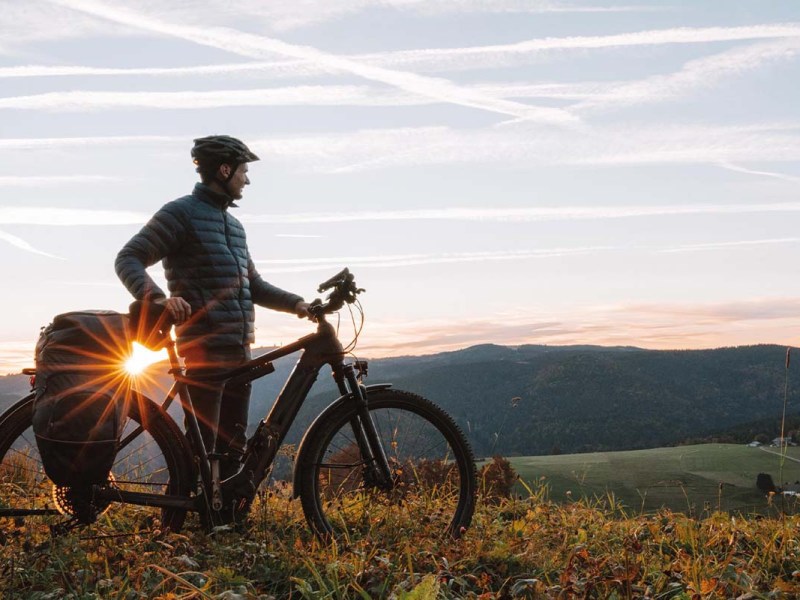 Mann mit seinem E-bike in der Natur auf einem Hügel, im Hintergrund die untergehende Sonne