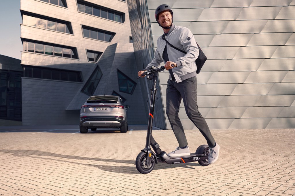 Mann fährt auf dem Audi electric kick scooter powered by Egret, im HIntergrund steht ein E-Auto von Audi