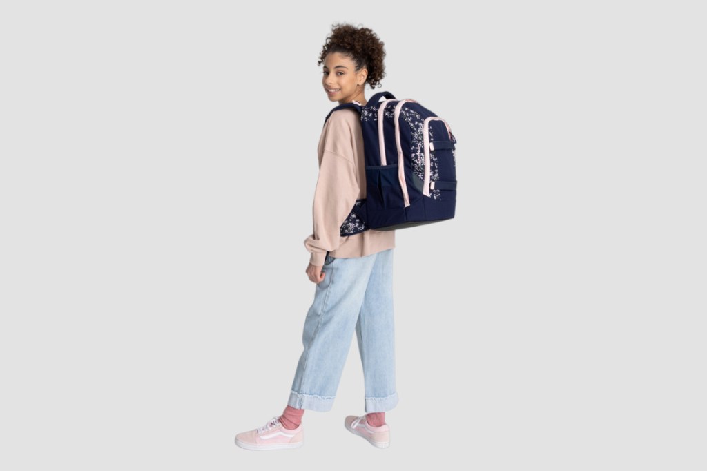Lächelndes Mädchen in rosa Pulli und blauer Hose schräg von hinten mit dunklem Satch Schulrucksack auf dem Rücken vor grauem Hintergrund