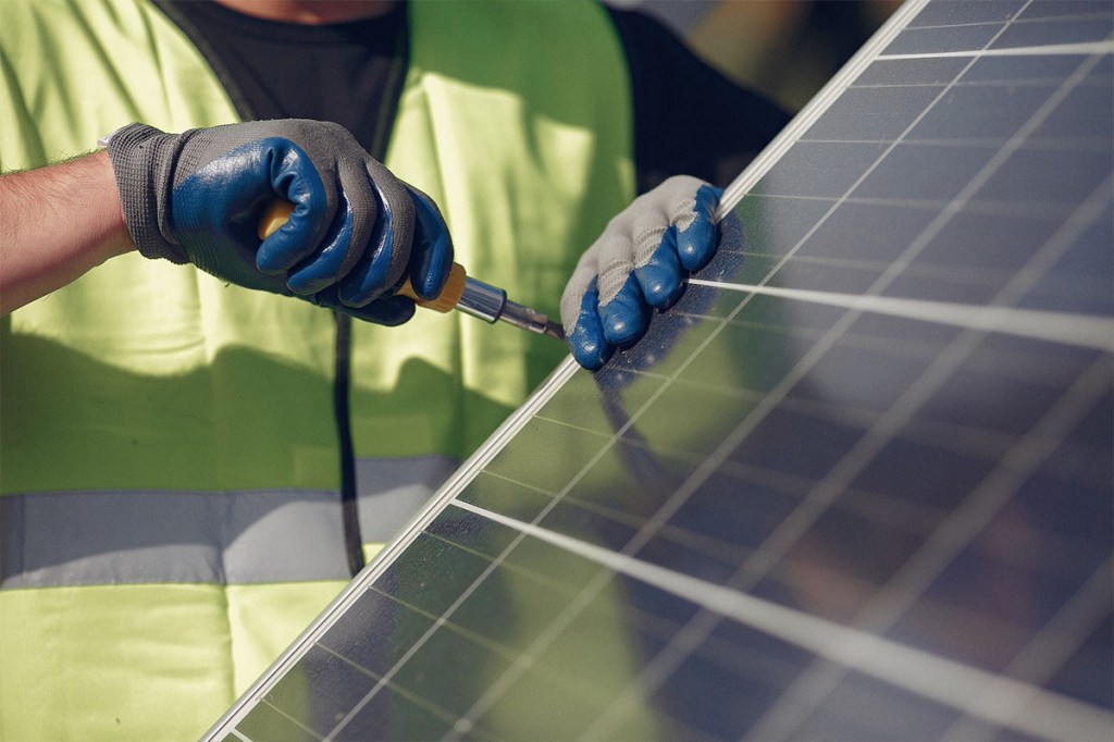 Eine Person mit Sicherheitsweste und Montage-Handschuhen schraubt an einem Solarpanel.