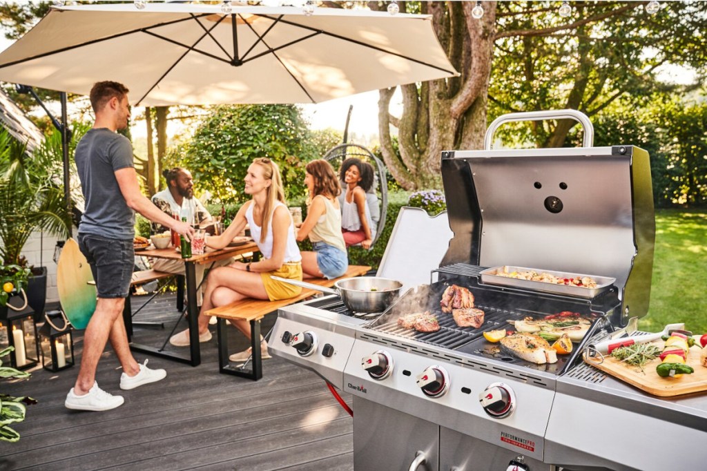 Rechts in der Ecke aufgeklappter silberner Gasgrill in Betrieb mit Fleisch und Gemüse am Braten, im Hintergrund Gruppe junger Menschen an Sitzgruppe unter Sonnenschirm auf Terrasse