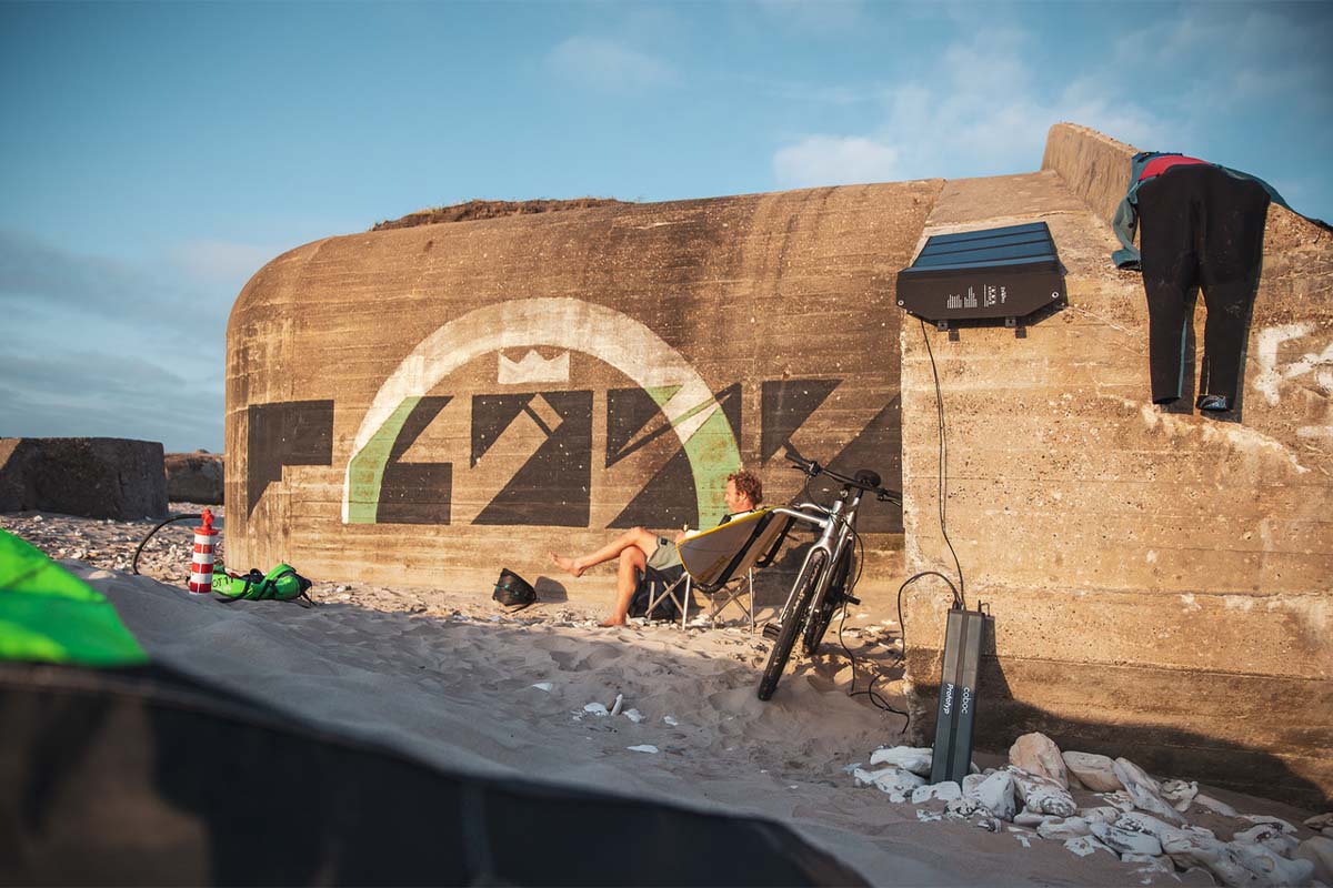 E-Bike Fahrer sitzt am Strand vor einer Mauer, neben im sein E-Bike, welches von einem Akku geladen wird