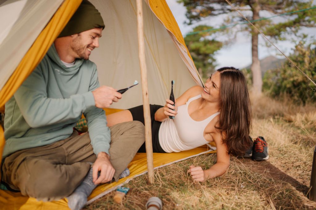 Ein Pärchen liegt in einem gelben Zelt in der Natur und putzt sich die Zähne mit der happybrush Eco VIBE 3 Schall-Zahnbürste.