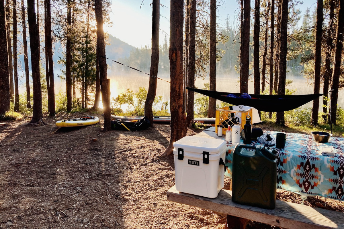 Waldlichtung bei Sonnenschein mit aufgebauten Campingtisch und Bänken auf dem Boxen und Trinkflaschen stehen, im Hintergrund eine Hängematte