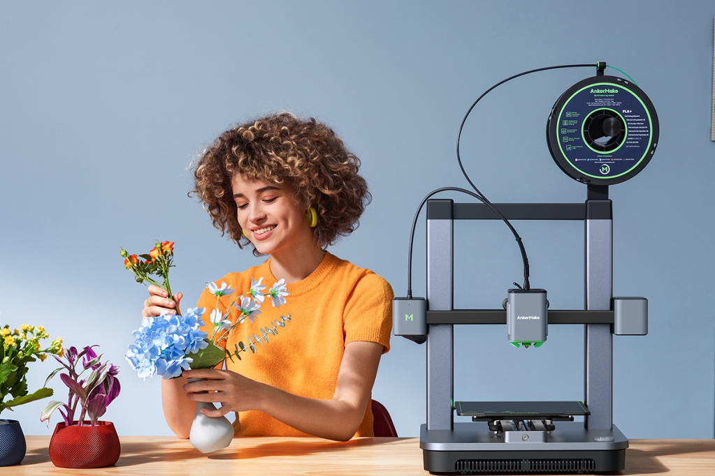 Eine Frau neben dem 3D-Drucker AnkerMake m5c in der der Hand eine gedruckte Vase mit einer Blume.