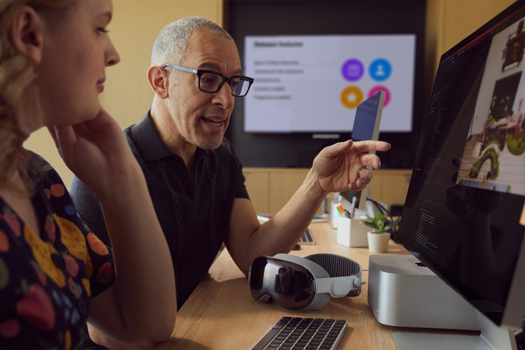 Zwei Menschen sitzen vor einem Computer, vor ihnen auf dem Tisch liegt die Apple Vision Pro-Brille.