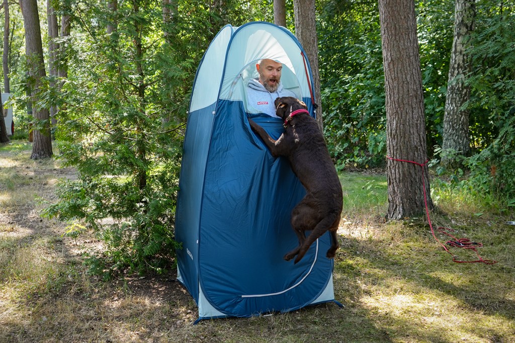 blaues Duschzelt im Wald stehend, Mann guckt mit dem Kopf aus der Tür heraus, Hund springt ihm von außen entgegen