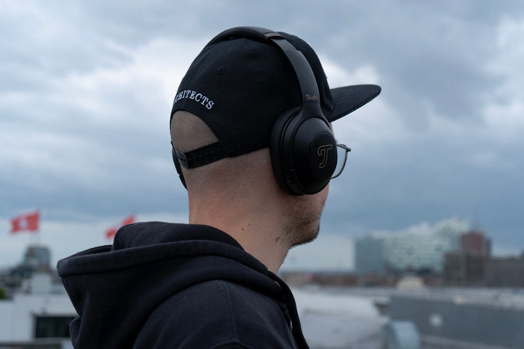 Real Blue Pro Kopfhörer von Teufel, getragen von einer Person mit Cap. Im Hintergrund Wolken und die Skyline von Hamburg.