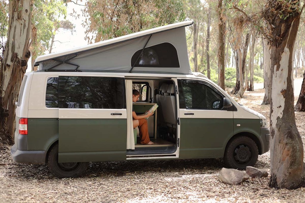 Camper in einem Wald stehend, die Tür ist geöffnet, man sieht ein Person im Anschnitt, die auf einer Campingtoilette sitzt