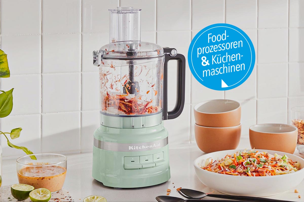 Mintgrüner zylinderförmiger Food Processor mit durchsichtigem Aufsatz in dem rotes Lebensmittel zerhackt ist auf weißem Tisch neben Teller und Schüsseln vor weißer Wand mit blauem Button rechts oben "Food-Prozessoren und Küchenmaschinen"