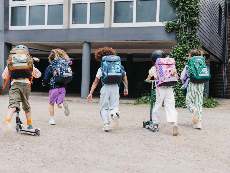 Fünf Kinder rennen in ein Schulgebäude, man sieht sie von hinten, alle tragen Ranzen