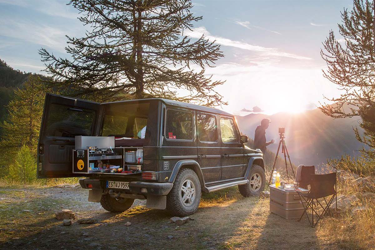 Auto steht in der Natur, Bergkulisse mit Sonnenaufgang im HIntergrund, man sieht eine Einbauküche im Heck des Autos