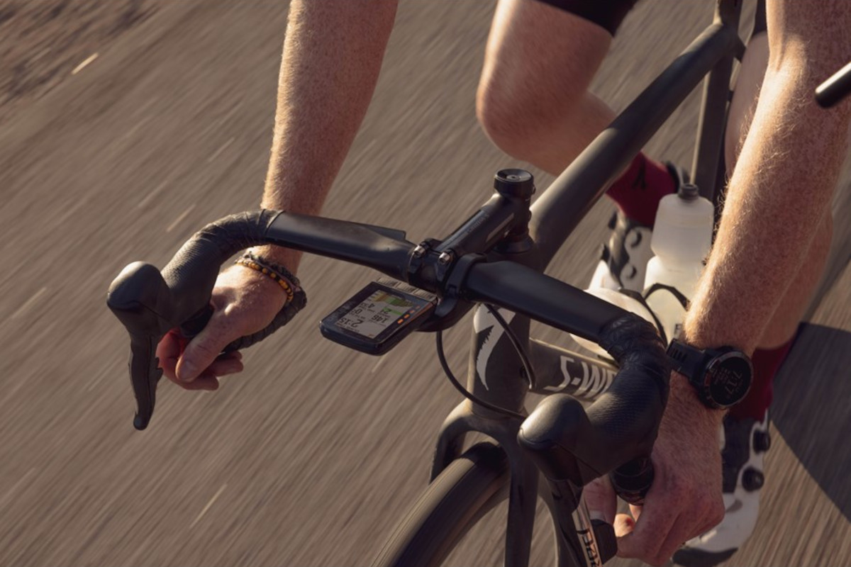 Schwarzes Rennrad fährt auf Asphalt und ragt von rechts oben ins Bild, Lenker wird von zwei Armen gehalten, am Lenker das rechteckige Fahrrad-Navi von Wahoo