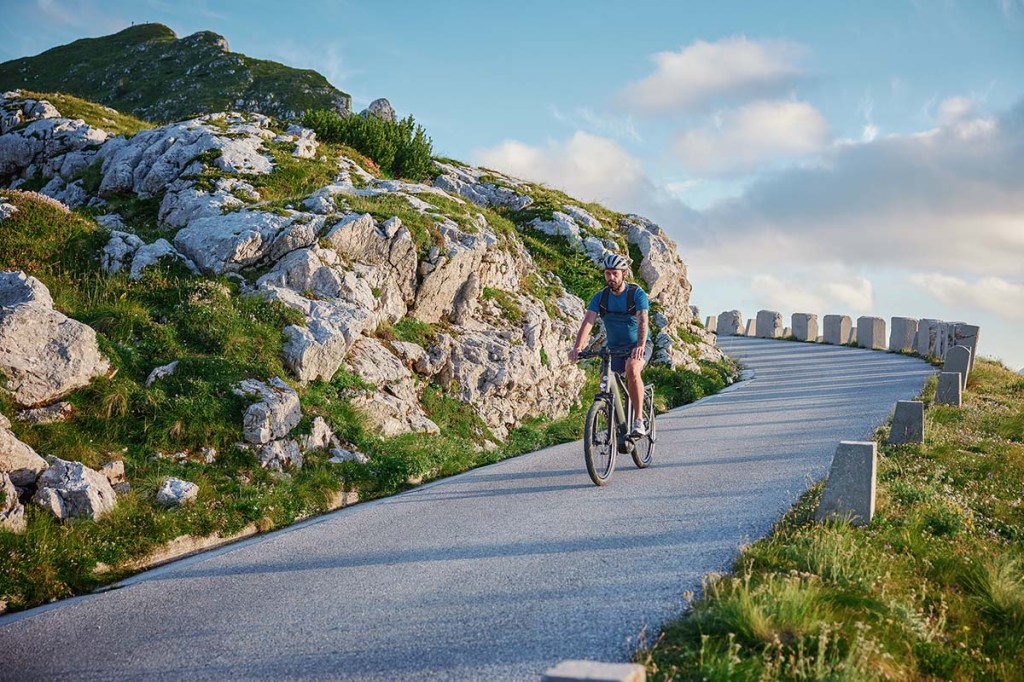 Mann fährt mit E-Bike eine Straße entlang, er ist umringt von einer Bergkulisse