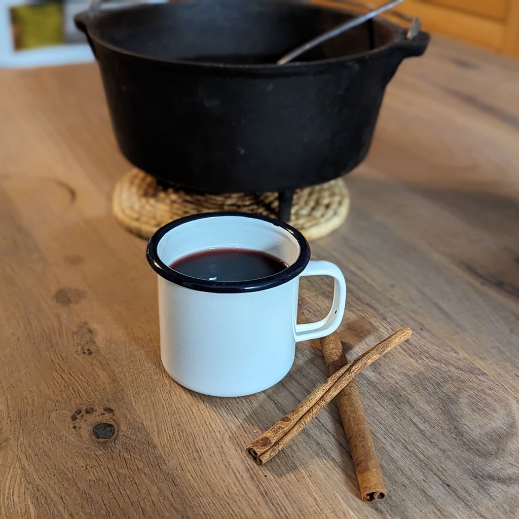 Eine Blechtasse mit Glühwein vor einem Dutch Oven. Daneben liegen zwei Zimtstangen