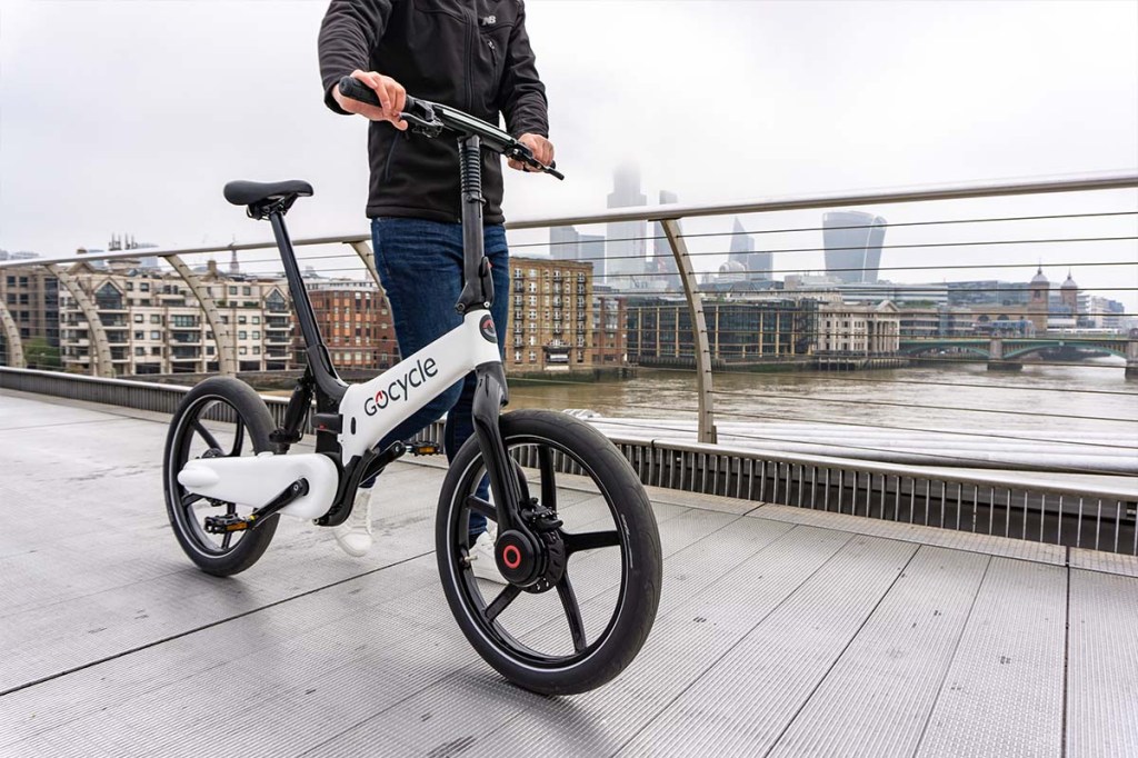 Person im Anschnitt, schiebt ein E-Bike über eine Brücke, Fluss mit Stadt-Skyline im Hintergrund