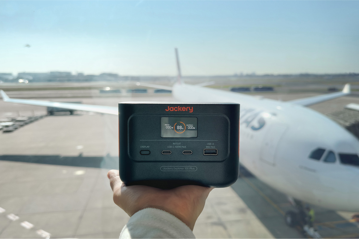 Eine ausgestreckte Hand, auf der die Jackery Explorer 100 Plus liegt. Dahinter ein Flugzeug am Flughafen.