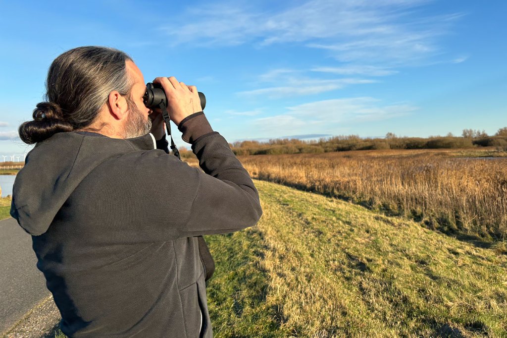 Ein Mann steht in der Natur und schaut mit dem Fernglas nach rechts.