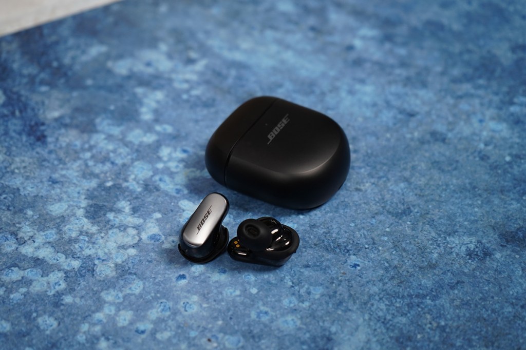 Die Quiet Comfort Ultra Earbuds neben ihrem schwarzen Ladecase liegend auf einem blauen Hintergrund fotografiert.