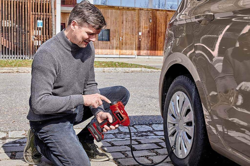 Mann kniet vor Auto und befüllt den Autoreifen mit einer Akku-Luftpumpe.