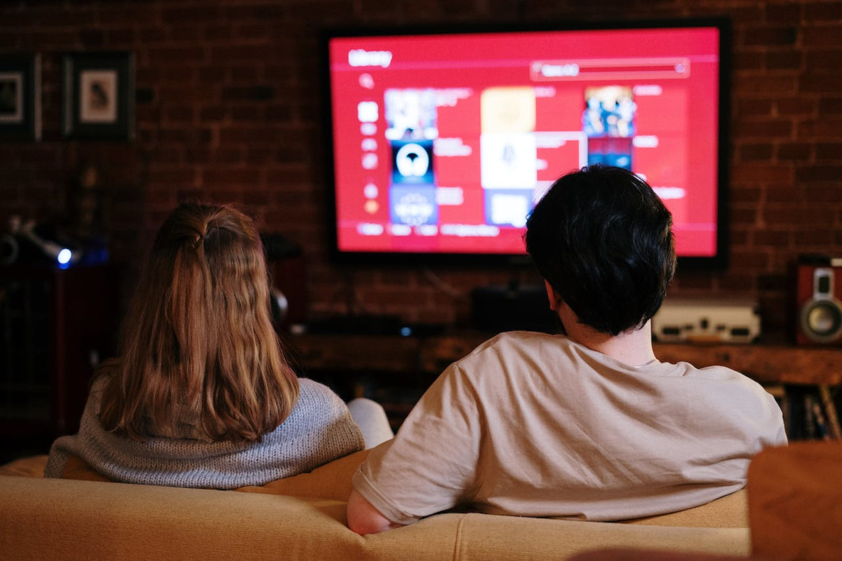 Frau von hinten mit langen hellbraunen Haaren sitzt zusammen mit Mann mit dunklen Haaren auf heller Couch vor Fernseher mit rotem Bildschirm