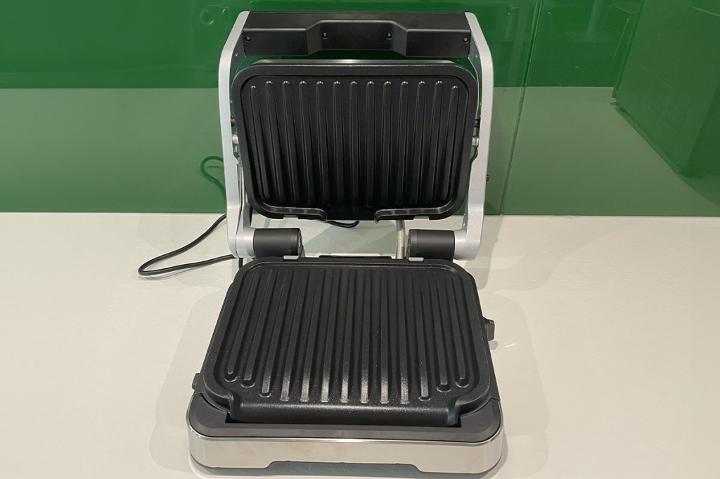 Der OptiGrill 4in1 mit geöffnetem Deckel auf einer weißen Arbeitsfläche vor einer grünen Wand.