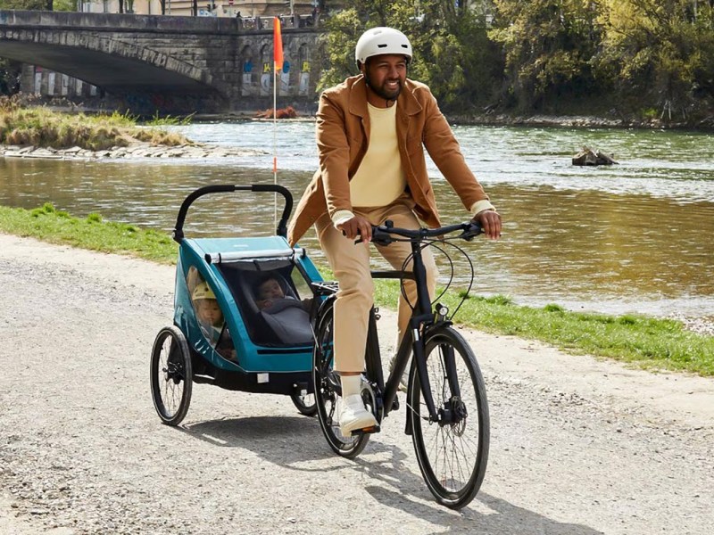 Mann auf einem Fahrrad mit Kinderanhänger an einem Fluss fahrend.