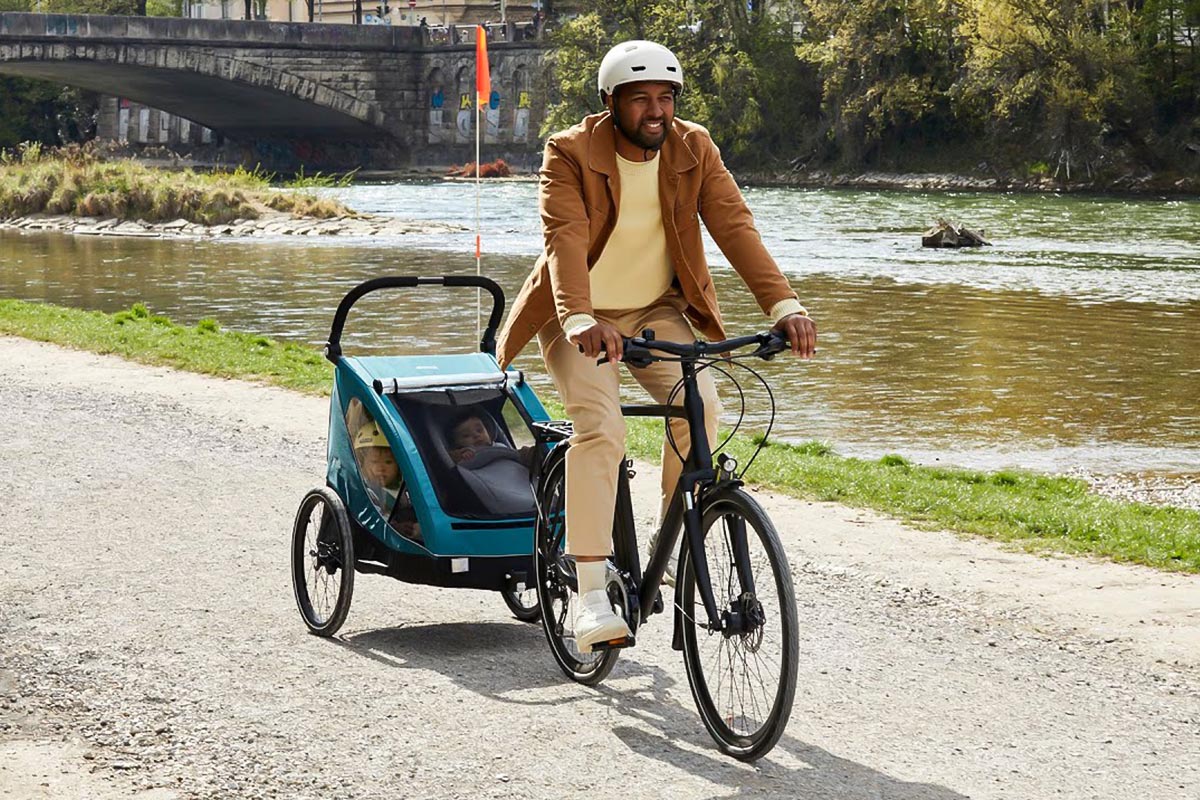 Mann auf einem Fahrrad mit Kinderanhänger an einem Fluss fahrend.