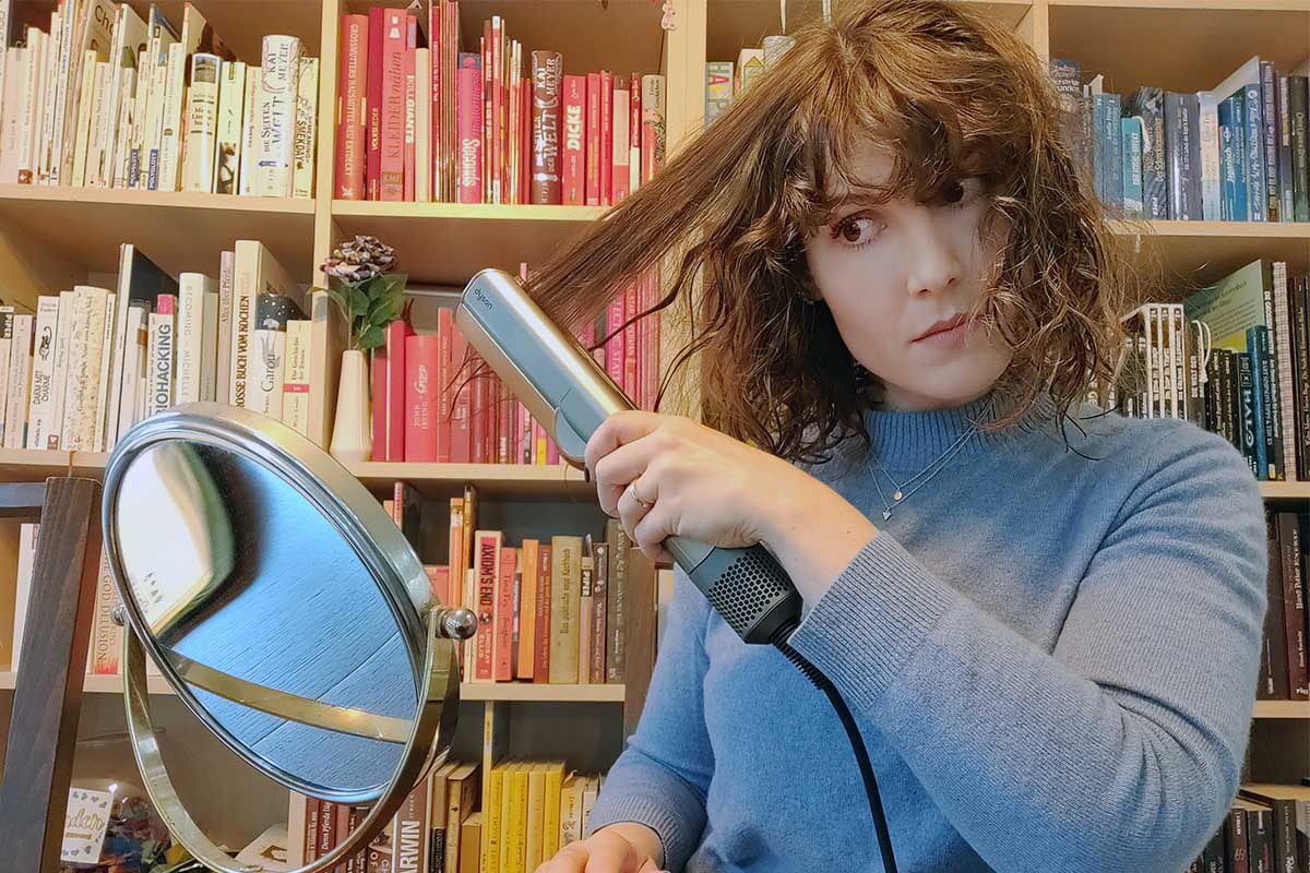 Die Testerin glättet eine Haarsträhne mit dem Dyson Airstrait. Hinter ihr ist eine bunte Bücherwand zu sehen.