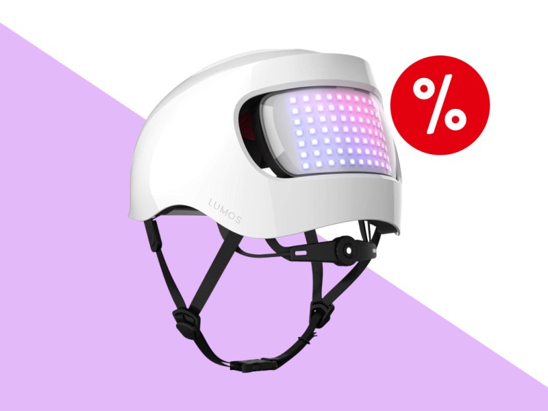 Weißer Lumos-Fahrradhelm schräg von hinten mit schwarzem hängenden geschlossenen Riemen und lila rosa leuchtender LED-Fläche auf lila weißem Hintergrund mit rotem Prozentbutton oben rechts