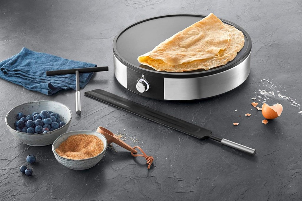 Der Crepe Maker WMF Lono auf einer schwarzen Tischplatte stehend. Daneben Küchenutensilien und Crepe-Zutaten.