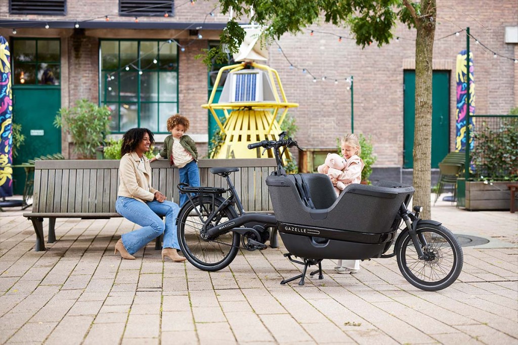 Zwei Frauen, eine mit Kiind, sitzen auf einer Bank in einem Innenhof, vor ihnen steht ein Cargo-E-Bike