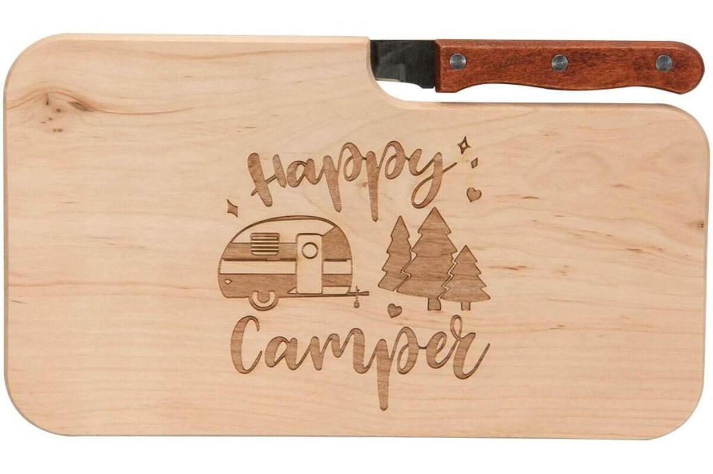 Helles rechteckiges Holzbrett mit Gravur von Wohnwagen und drei Tannen sowie dem Spruch Happy Camper, mit Messer mit dunklem Holzgriff oben eingesteckt auf weißem Hintergrund