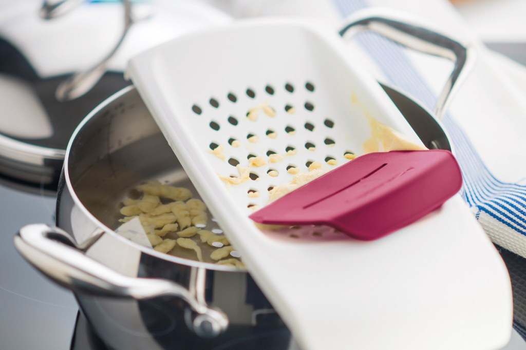 Weiße Reibe über silbernen Kochtopf in dem Wasser mit Spätzle sind, darauf ein roter Hobel