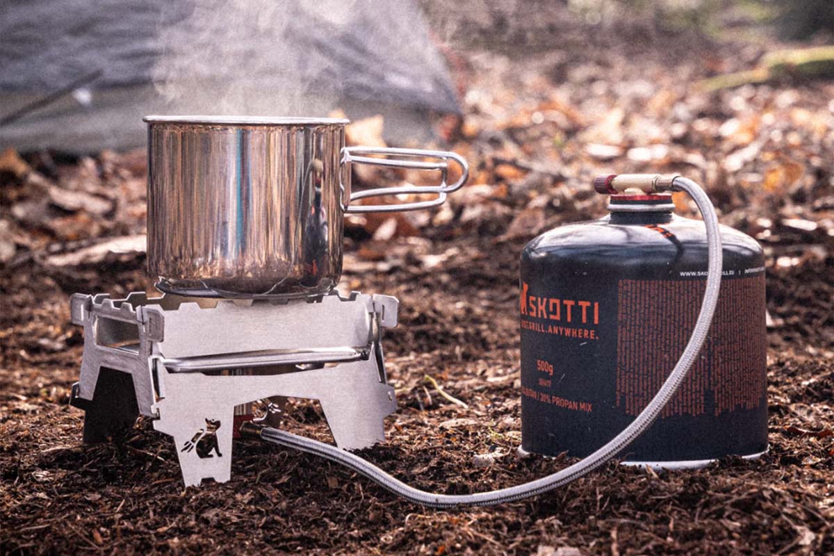 Kleiner Gaskocher auf einem Waldboden stehend, auf dem ein Topf steht. Er ist an eine Gaskartusche angeschlossen