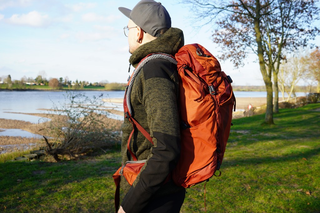 Mann von der SEite in der NAtur stehend, er hat einen roten Rucksack auf dem Rücken