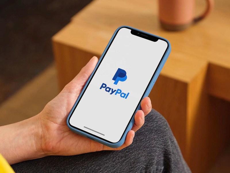Eine Person hält ein Smartphone in der Hand, auf dem das Paypal Logo zu sehen ist.