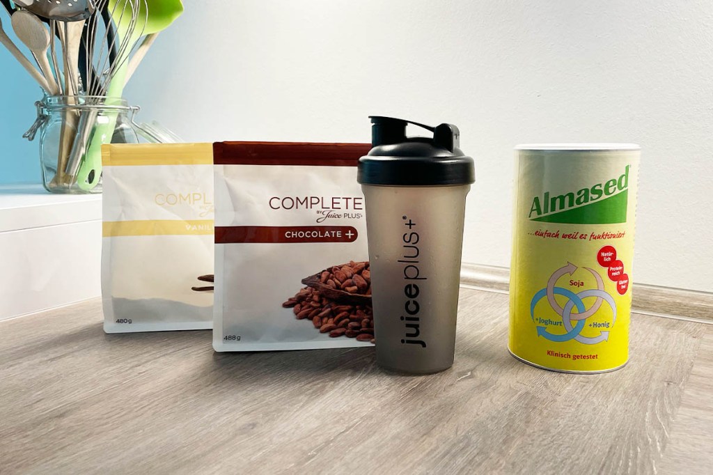 Zwei Packungen JuicePlus+ Complete Pulver, ein Shaker und eine Packung Almased auf einer Küchenarbeitsplatte.